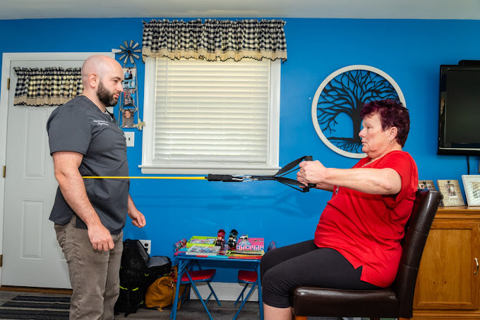 Physical therapist guiding a seated patient through a resistance band exercise at home