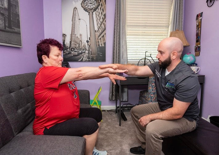 Physical therapist guiding a seated patient through an arm-reaching exercise at home