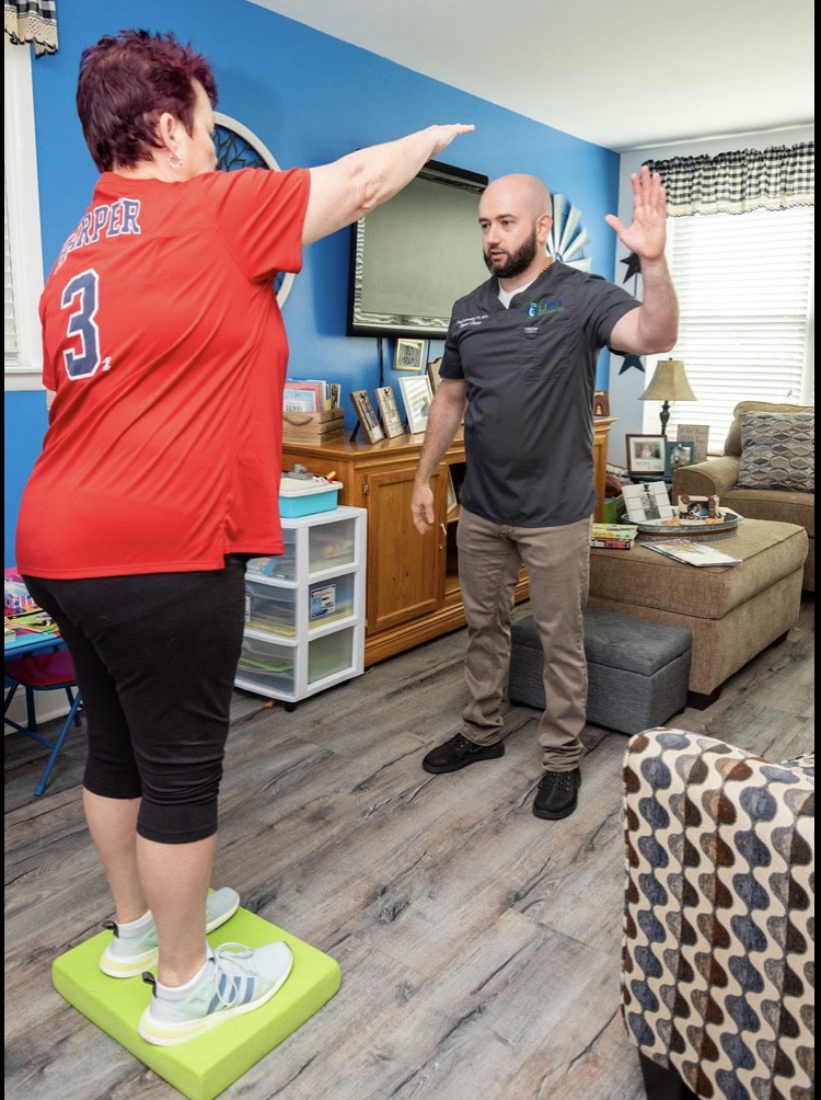 Patient standing on a foam balance pad and reaching overhead as a physical therapist demonstrates the exercise